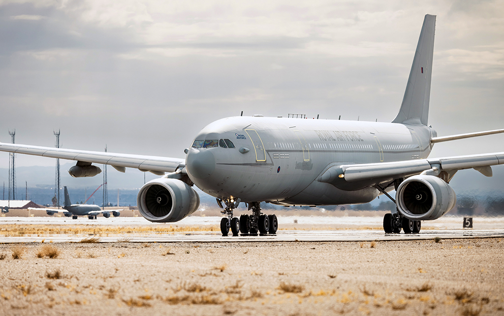 A Royal Air Force Voyager transport aircraft, sporting the RAF livery, taxis on a desert runway under a cloudy sky. Another military aircraft is visible parked in the distant background.